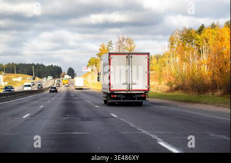 Camion de livraison blanc de vue arrière sur la route asphaltée de l'autoroute à plusieurs voies, flux de trafic avec des voitures et des fourgonnettes, arbres d'automne vibrants le long de la route, nuageux Banque D'Images