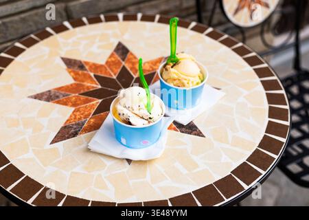 Gros plan de cuillères à crème glacée appétissantes dans des gobelets en papier reposant sur une table en mosaïque colorée dans un café en plein air. Texture détaillée du dessert glacé et vi Banque D'Images