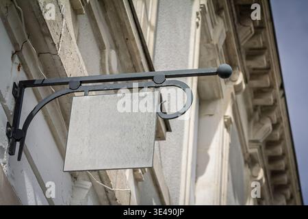 Panneau suspendu carré gris vide monté sur une façade extérieure ornée, gros plan avec une faible profondeur de champ. Panneau de signalisation vierge pour la marque du magasin, copie Banque D'Images