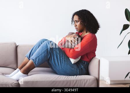 Jeune femme est assise sur le canapé tenant un oreiller et regardant réfléchi dans une salle lumineuse Banque D'Images
