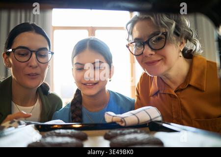 Femmes d'âge moyen avec une fille vérifiant les cookies dans le four, cuisant ensemble à la maison et partageant l'activité familiale. Utile pour les parents, les générations, la cuisine à la maison, le marketing de style de vie Banque D'Images