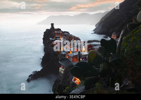 Vue sur le village illuminé de Vernazza perché sur une falaise rocheuse surplombant la mer au crépuscule avec des cactus de poire épineuse au premier plan Vernazz Banque D'Images