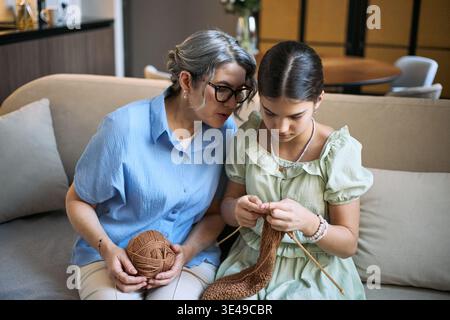 Femme senior guidant la fille tout en tricotant avec du fil sur le canapé, partageant passe-temps fait main et apprentissage intergénérationnel. Utile pour les liens familiaux, l'éducation artisanale, le marketing des activités à domicile Banque D'Images
