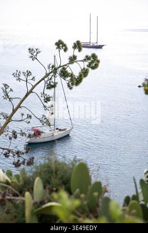 Vue d'un voilier blanc ancré dans les eaux bleues calmes de la mer Méditerranée encadrée par la végétation côtière à Lipari, Sicile, Italie. Banque D'Images