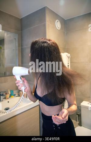 Femme se tient dans la salle de bain moderne sèche de longs cheveux foncés avec un sèche-cheveux, routine de soins personnels du matin décontractée dans une lumière naturelle douce, authentique, indépendant Banque D'Images