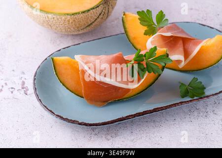 Tranches de jambon espagnol traditionnel (viande de porc séchée) avec melon (cucumis melo) et feuilles de persil frais herbes sur assiette vintage. Plat sur table de pierre légère Banque D'Images