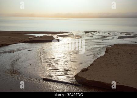 Ruisseaux de marée peu profonds coulant sur la plage de sable dans la mer avec des reflets lisses sous un ciel brumeux doux. Banque D'Images