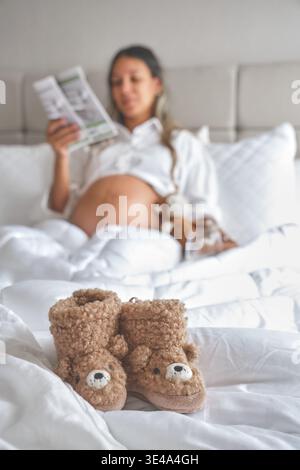 Femme enceinte lisant le journal au lit avec des bottines de bébé au premier plan Banque D'Images