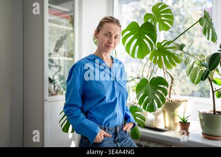 Joyeuse femme d'âge moyen heureuse appréciant le jardinage en intérieur, posant près de plantes en pot au rebord de la fenêtre. Banque D'Images