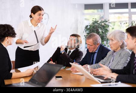 Intelligent jeune femme faisant rapport tandis que les employés de bureau d'âges différents utilisant des cahiers à la table dans le bureau Banque D'Images