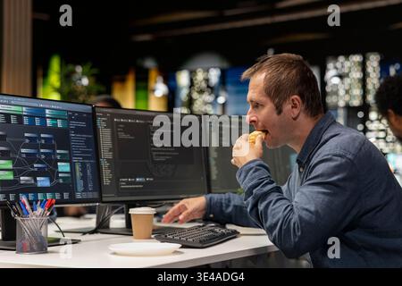 Développeur de logiciels mangeant croissant au bureau, examinant les métriques du système d'IA, surveillant les tableaux de bord de performance. Expert EN INFORMATIQUE DE LA société de technologie profitant d'une pause collation au bureau tout en utilisant le programme sur l'ordinateur Banque D'Images