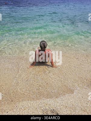 Femme en bikini assise dans une eau turquoise peu profonde sur une plage de sable, vue arrière, appuyée sur les mains et regardant la mer Ionienne à Nikiana, Lefkada. Banque D'Images