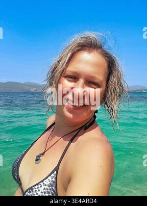 Femme caucasienne souriante en bikini à motifs faisant un portrait selfie en gros plan avec la belle mer Ionienne et les collines verdoyantes à Nikiana, Lefkada, Grèce. Banque D'Images