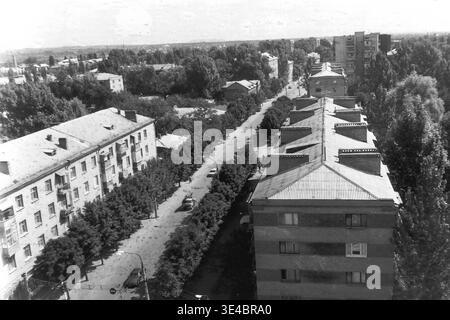 Slavyansk, 1998, rue Yunykh Kommunarov (aujourd'hui Bankivska). Archive photographie en noir et blanc présentant une belle vue panoramique surélevée depuis un bâtiment de neuf étages sur une avenue résidentielle tranquille. Le paysage urbain présente des blocs d'appartements en briques classiques à plusieurs étages parfaitement bordés d'arbres d'été denses et matures, créant une atmosphère de quartier calme, avec des cheminées détaillées sur le toit au premier plan. Ce paysage urbain capture merveilleusement l'environnement architectural et l'ère d'un Donbass paisible. Banque D'Images