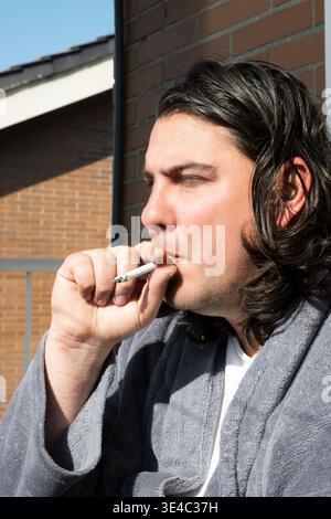 Homme aux cheveux longs fumant une cigarette tout en portant un peignoir à l'extérieur sur un balcon Banque D'Images