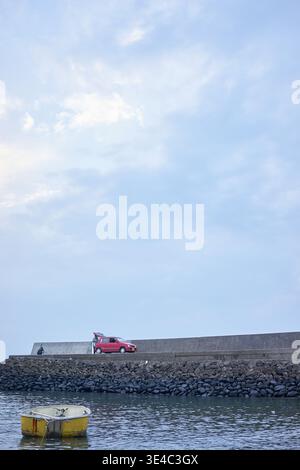 Un petit port serein avec un bateau jaune flottant, une voiture rouge, et les gens profitant d'une journée de détente de pêche sous un ciel calme et couvert. Banque D'Images
