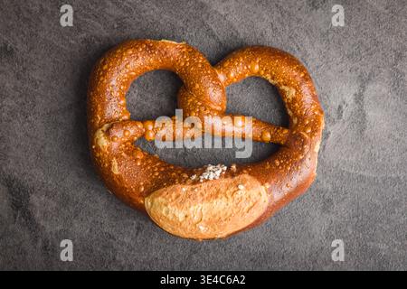 Bretzel doux salé brun doré photographié de haut en bas sur une surface texturée sombre. Frais d'une boulangerie artisanale, le pain torsadé montre du gros sel, Banque D'Images