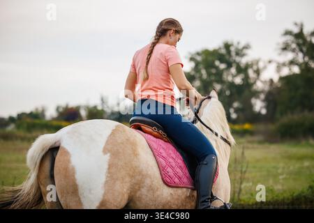 Femme à cheval sur la prairie. Activité de loisirs équestres au ranch en campagne. Vue arrière Banque D'Images