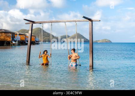 Un couple diversifié se détend sur des balançoires dans les eaux peu profondes de Sainte-Lucie, entouré de montagnes luxuriantes et d'un ciel bleu vif. Ce paradis tropical offre l'escapade parfaite pour des vacances de luxe Banque D'Images