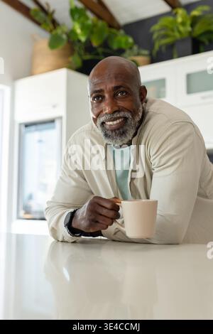Homme afro-américain senior dans un cardigan beige appuyé sur l'île blanche dans la cuisine tenant une tasse beige Banque D'Images