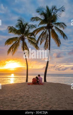 Un couple est assis ensemble sur une plage de sable sous les palmiers, partageant un moment de calme alors que le soleil se couche sur la mer des Caraïbes. Cette scène idyllique capture l'essence du romantisme à Sainte-Lucie. Banque D'Images