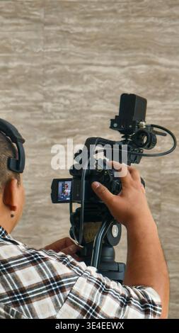 Personne utilisant une caméra vidéo professionnelle sur trépied, portant une chemise à carreaux et un casque, ajustant les paramètres de la caméra à l'intérieur. Banque D'Images