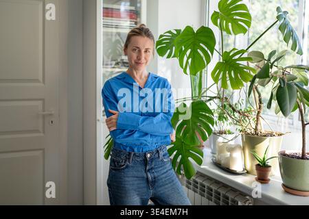 Positive femme d'âge moyen amant de plantes posant à côté de Monstera en pot dans le jardin de la maison avec rebord de fenêtre. Banque D'Images