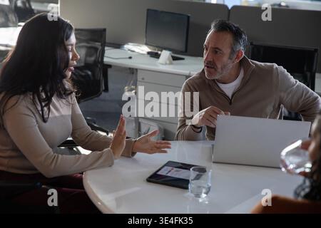 Divers collègues discutant, assis à une table ronde blanche avec un ordinateur portable en argent, une tablette, un verre d'eau Banque D'Images