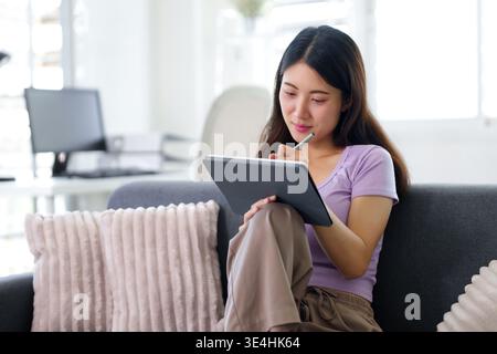 La femme adulte asiatique focalisée utilise un stylet sur tablette. Elle travaille à distance à la création de projets en apprenant à partir de l'environnement moderne de bureau à domicile. Banque D'Images
