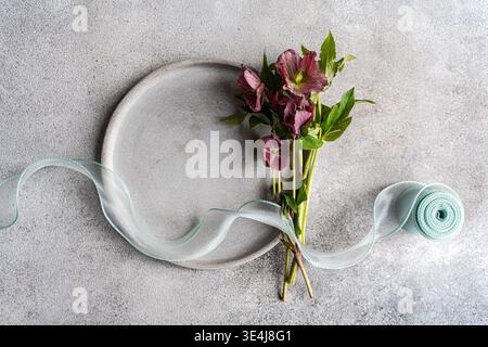 Pose plate minimale de fleurs de printemps hellebore avec des feuilles vertes à côté d'une plaque vierge, stylisée avec un ruban pâle sur un fond de pierre grise, idéal pour e. Banque D'Images