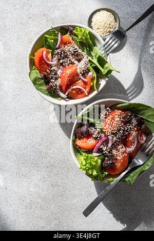 Vue de dessus de deux bols de salade d'épinards frais avec des tranches de tomate, des olives noires, de l'oignon rouge et une généreuse garniture de graines de sésame sur une surface de pierre légère. Banque D'Images