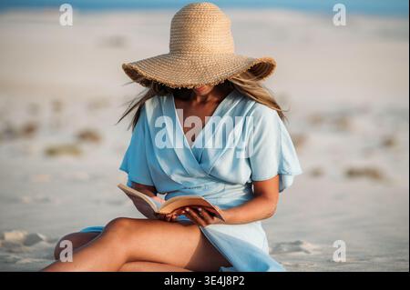 Femme détendue dans un chapeau de soleil lit un livre tout en étant assise sur la plage de sable à Corralejo, Fuerteventura, Espagne, profitant d'un moment de vacances d'été calme par Banque D'Images