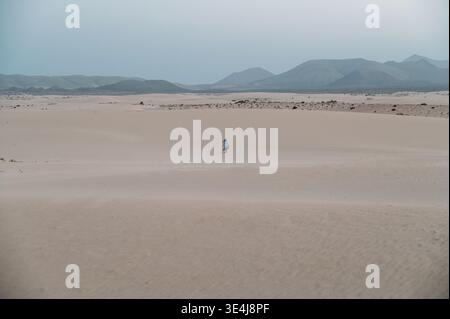 Une femme solitaire marche à travers de vastes dunes de sable à Corralejo, Fuerteventura, Espagne, avec des montagnes brumeuses à l'horizon sous un ciel couvert, minimalis Banque D'Images