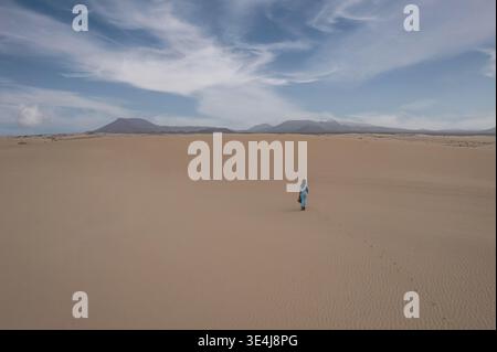 Femme solitaire marche à travers de vastes dunes désertiques à Corralejo, Fuerteventura, Espagne. Paysage minimaliste avec sable ondulé, montagnes lointaines et drame Banque D'Images