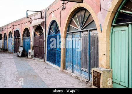 Vieilles portes marocaines en bois vieilles vieilles vieilles vieilles portes en bois sous des façades voûtées bordent une rue calme d'Essaouira, avec des bleus et des pastels délavés, du plâtre texturé et du histo Banque D'Images