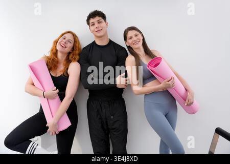 Trois amis se tiennent ensemble, souriants et détendus, tenant des tapis de yoga roses. Ils sont vêtus de vêtements de sport, incarnant un mode de vie sain et actif Banque D'Images