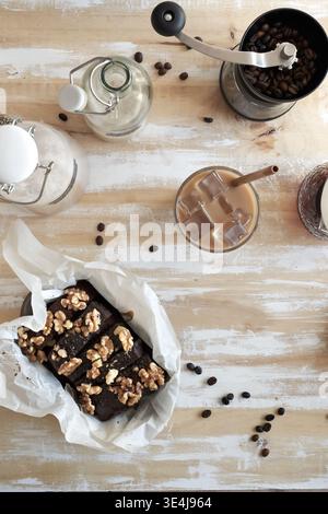 Pose à plat de café glacé avec de la paille à côté d'un moulin manuel, grains de café, bouteille en verre et pot, plus brownie au chocolat et aux noix sur bois rustique Banque D'Images