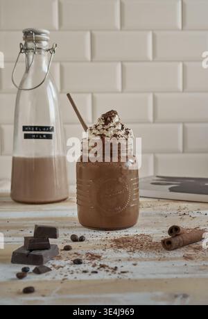Milkshake au chocolat rustique dans un pot mason garni de crème fouettée et de cacao, avec une bouteille de lait aux noisettes, des morceaux de chocolat, des grains de café et de la cannelle Banque D'Images