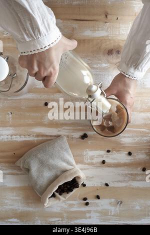 Vue de dessus des mains versant du lait d'une bouteille en verre dans du café glacé dans un pot sur une table en bois rustique, avec des grains de café dispersés et une pochette de lin, Banque D'Images