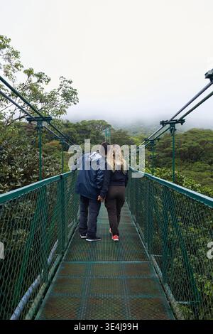 Un couple marche main dans la main sur un pont suspendu immergé dans la verdure luxuriante de la forêt tropicale de Monteverde au Costa Rica, entouré de nuages brumeux Banque D'Images