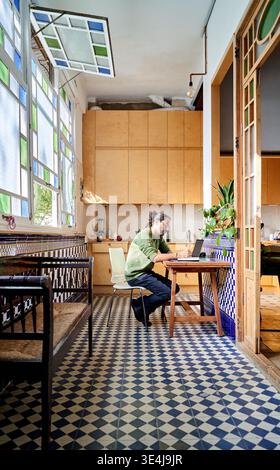 Un homme de descendance hispanique travaille à une table en bois dans une cuisine lumineuse avec des carreaux colorés. Il est concentré sur son ordinateur portable lorsqu'il est assis sur une chaise. Banque D'Images