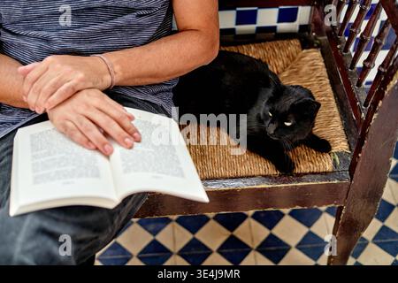 Une personne de descendance hispanique est assise sur un banc à la maison avec un livre à la main. Un chat noir se repose à côté d'eux. Cette scène montre un travail relaxant de la maison Mo Banque D'Images