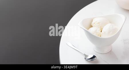 La crème glacée à la vanille est servie dans un plat en céramique blanche en forme de bateau, avec trois généreuses cuillères visibles. Une cuillère en métal brillant repose sur le blanc, subtilement mar Banque D'Images