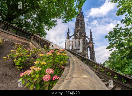 Vue spectaculaire en bas angle du Scott Monument encadré par des arbres feuillus, des escaliers en pierre et des hortensias en fleurs. L'architecture gothique historique contraste avec Banque D'Images