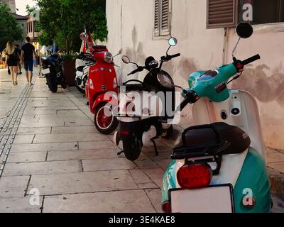 Une rangée vibrante de scooters à moteur en rouge, blanc et vert menthe stationné le long d'un trottoir en pierre légère. L'arrière-plan montre une scène de rue floue avec pe Banque D'Images