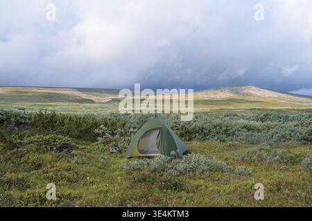 Une photographie panoramique grand-angle capturant une tente à dôme vert solitaire pour le backpack niché au milieu de la végétation basse sur une vaste toundra arctique ondulée Banque D'Images
