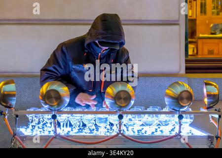 Interprète de rue à capuche joue un instrument en verre fait de verres à vin sous des lumières vives, performance musicale créative en direct la nuit. Banque D'Images