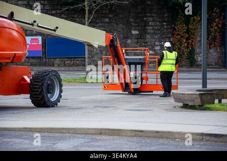 Travailleur de la construction en gilet réfléchissant et casque de sécurité debout à côté d'un ascenseur orange boom dans une rue de la ville. Banque D'Images