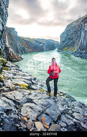 Un randonneur se tient sur les rives rocheuses du canyon Studlagil, entouré d'imposantes colonnes de basalte et d'eaux vertes coulantes sous un ciel nuageux en Islande. Banque D'Images