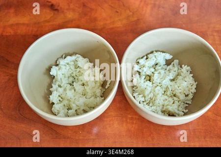 Deux bols identiques en céramique beige clair contenant des portions de riz blanc cuit, placés côte à côte sur une surface de table chaude en bois brun rougeâtre. Banque D'Images